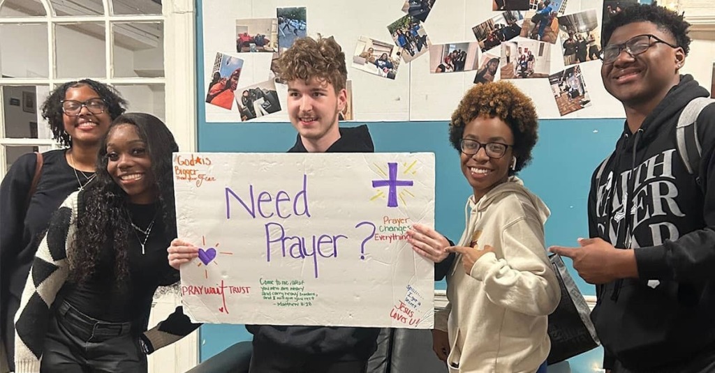 Five student members of Bethel Campus Fellowship at Virginia State University hold up a sign illustrated with a cross and the question, "Need Prayer?"