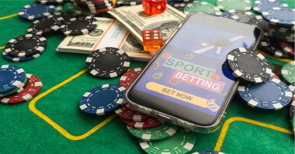 A photo showing money, poker chips, and smartphone opened to an online sports gambling platform that reads "Bet Now."
