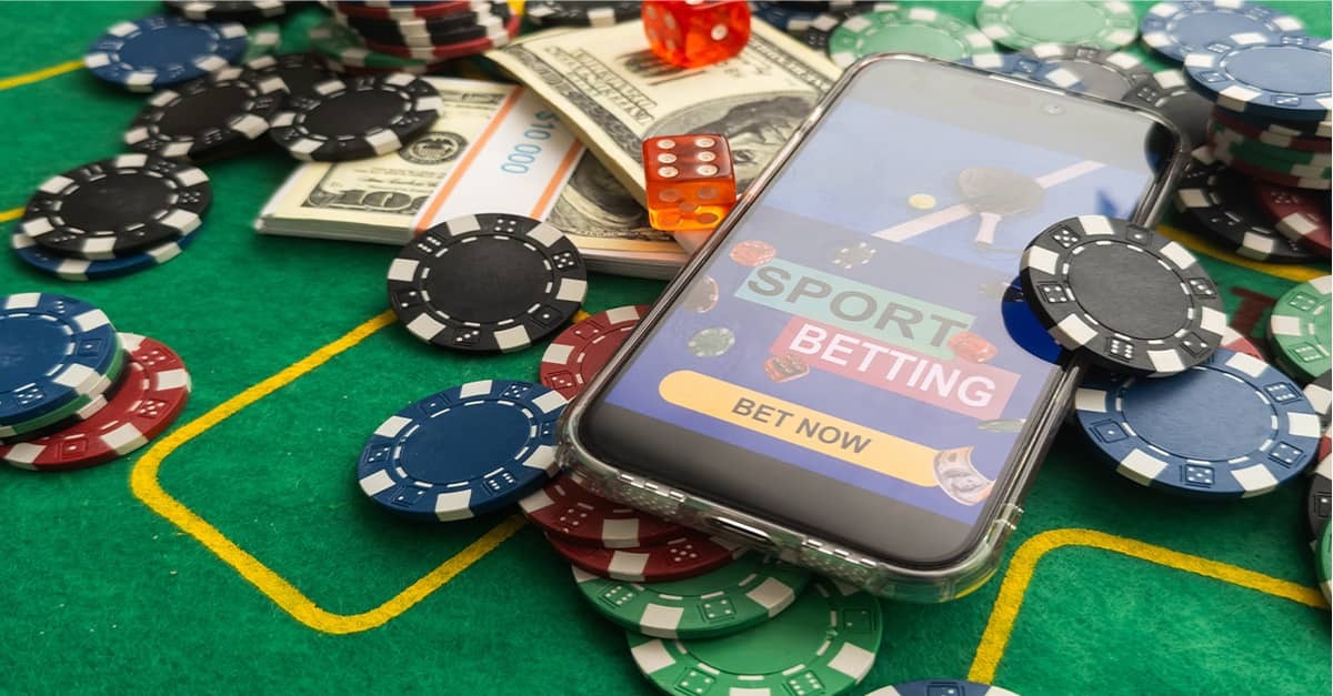 A photo showing money, poker chips, and smartphone opened to an online sports gambling platform that reads "Bet Now."