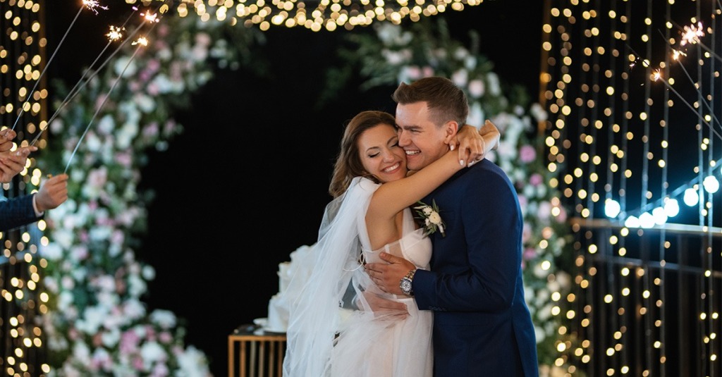 A happy bride hugs her new husband around the neck against a backdrop of lights and sparklers.